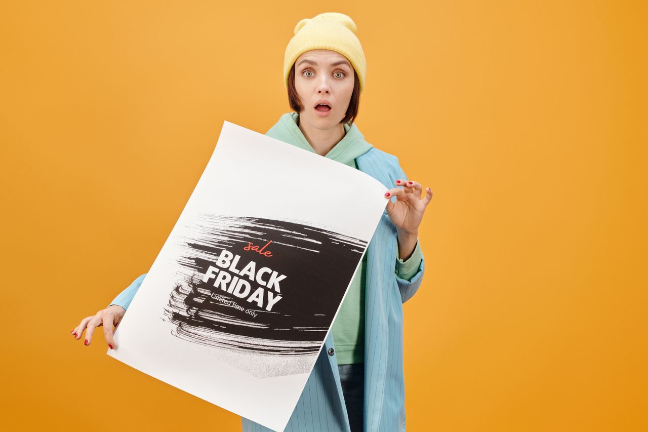 services-04 Young woman holds a Black Friday sale poster, surprised against a yellow background.